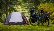 Grå og orange kuppeltelt opstillet i en skov ved siden af en touringcykel med flere tasker, ideel til udendørs eventyr og cykelture med oppakning.