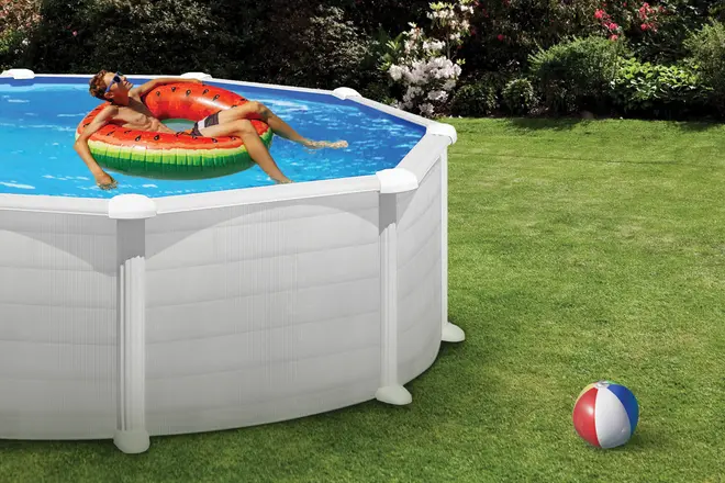 Rund fritstående pool med hvid yderside, fyldt med vand. Inkluderer en farverig badering. Poolen står på grøn græsplæne med en strandbold i nærheden.
