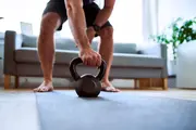 Sort kettlebell på en yogamåtte, grebet af en person i hjemmetræning med sofa og plante i baggrunden.