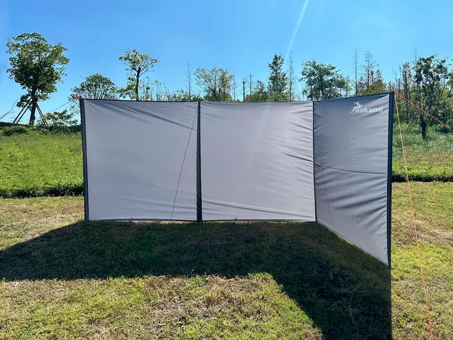 Udendørs vindskærm med tre sammenhængende grå stofpaneler, støttet af stænger og barduner. Giver læ og privatliv til camping eller picnic. Let og nem at opstille.