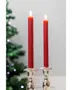 To høje, røde stagelys er tændt og placeret i elegante sølvlysestager, perfekte til festlige eller formelle borddækninger. Sløret juletræ i baggrunden.