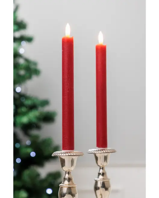 To høje, røde stagelys er tændt og placeret i elegante sølvlysestager, perfekte til festlige eller formelle borddækninger. Sløret juletræ i baggrunden.