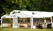 Stor hvid festtelt med gennemsigtige sider, opstillet udendørs på græs. Indeni er dekorerede borde og stole, ideelt til bryllupper eller fester. Elegant og rummeligt.