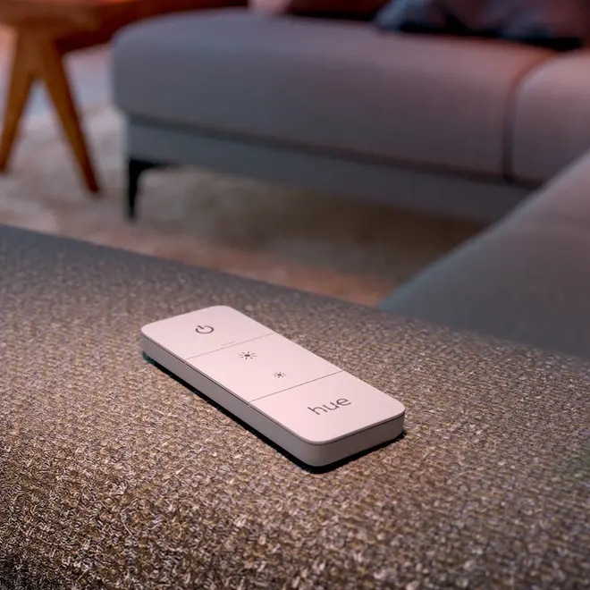 Hvid Philips Hue fjernbetjening med fire knapper: tænd/sluk, lysstyrke op, lysstyrke ned og hue. Slankt, rektangulært design, placeret på stofoverflade.