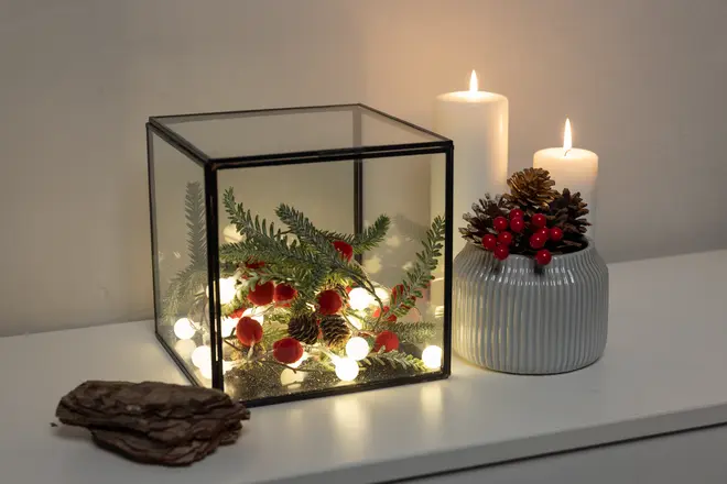 Glaskube med grangrene, røde bær og lyskæde. Ved siden af står en rillet vase med kogler og bær samt to tændte hvide stearinlys. Dekorative træstykker medfølger.