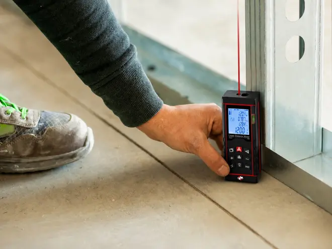 Kompakt laserafstandsmåler med oplyst display, knapper og rød laserlinje, holdt mod en væg til præcis gulv-til-loft-måling.