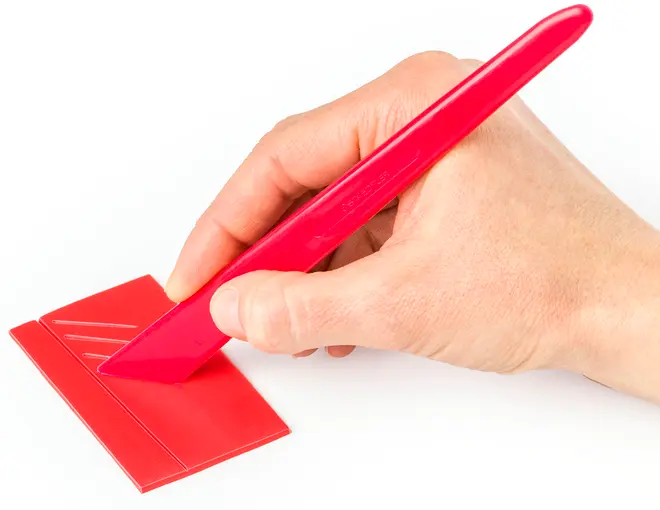 Rødt plastfoldningsværktøj med ergonomisk håndtag, bruges til at lave præcise folder i papir eller karton. Vist i brug på et rødt ark med prægede linjer.