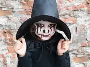 Børns Halloween-kostume med sort heksehat, sort kappe og ansigtsmaling med hvid base, sorte og røde detaljer samt syet mund. Perfekt til uhyggelige fester.