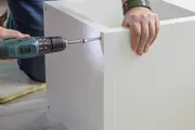 Person monterar ett vitt, rektangulärt plattpaket-skåp med en borrmaskin som skruvar i sidopanelen. Skåpet har en slät, modern yta.