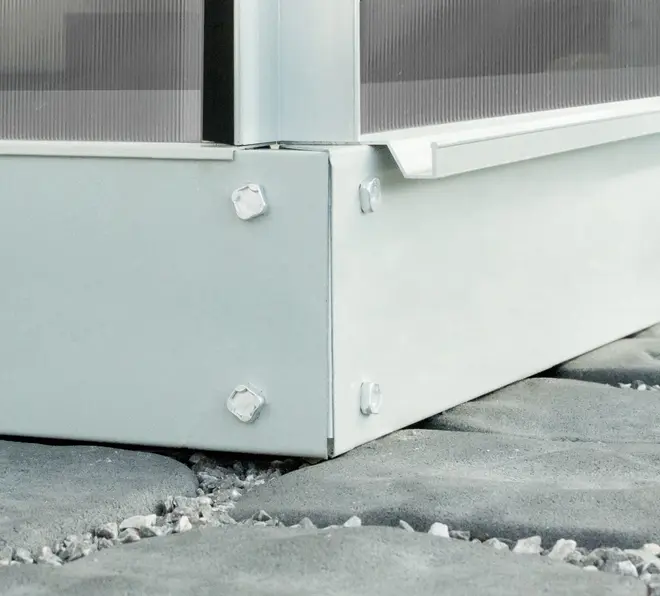 Närbild av växthusets metallhörn med synliga bultar, placerat på stenplattor. Konstruktionen har en robust, galvaniserad stålram och polykarbonatpaneler.