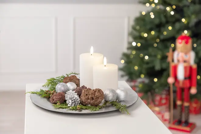 Dekorativt fat med två vita blockljus, silverkulor, kottar, bruna stenar och gröna kvistar, arrangerat som juldekoration. Julgran och nötknäppare i bakgrunden.