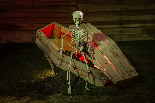Halloween-dekoration med ett skelett i naturlig storlek sittande i en delvis öppen träkista med rött ljus, placerad på konstgräs, perfekt för läskig utomhusdekoration.