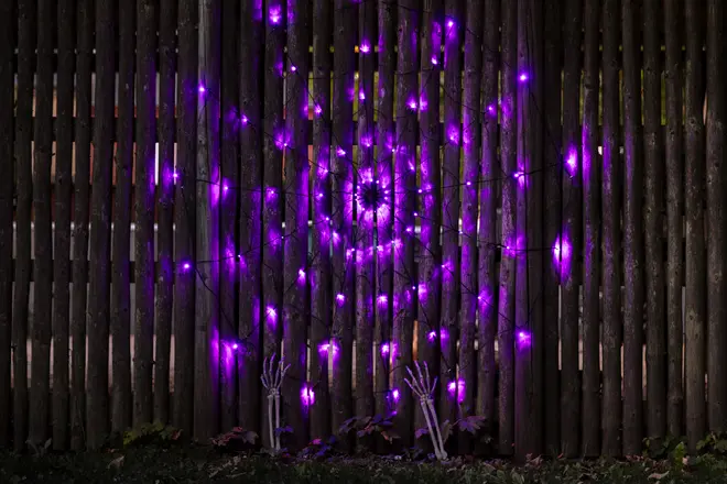 Halloween-dekoration med lila LED-spindelnätsljus på ett trästaket och två plast-skelett händer längst ner för en kuslig utomhusdekoration.