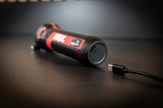 Rød og sort cylindrisk genopladelig enhed med USB-C-ladeport, sidepanel med display og tilsluttet ladekabel på mørkt bord.