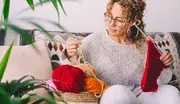 Kvinde strikker med rødt garn, holder strikkepinde og en kurv med rødt, orange og pink garn, sidder på mønstret sofa iført lysegrå sweater.