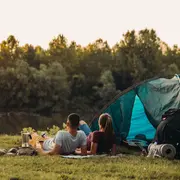 Blå kuppeltelt med åben indgang, opstillet på græs nær en sø. Inkluderer sovemåtte, rygsæk og kedel. Passer til to personer, udendørs brug.