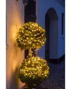 Udendørs lyskæde snoet omkring to kugleformede topiary-buske, udsender varmt hvidt lys. Perfekt til have- eller terrasseudsmykning og skaber en hyggelig stemning om aftenen.