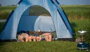 Blå kuppeltelt på græs med åben front, hvor fire personer ligger indeni. Udendørs står et bærbart gasblus, perfekt til camping og familieudflugter.