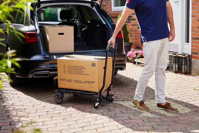 Sammenklappelig sort sækkevogn med fire hjul, bærer en stor papkasse. Kompakt design, ideel til flyttekasser eller bagage. Vist udendørs ved bil og husindgang.