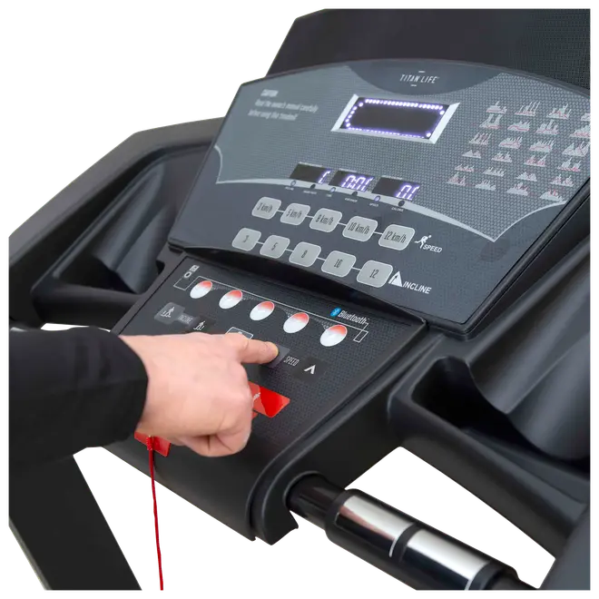 Nærbillede af løbebåndets kontrolpanel med digitalt display, flere hastigheds- og hældningsknapper, Bluetooth-forbindelse og en person, der trykker på en rød knap. Sort stel med kopholder.