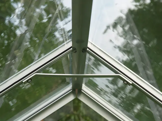 Närbild av ett växthustak med klara glasrutor, aluminiumram och stödstång. Konstruktionen är modern, stabil och släpper in maximalt med ljus till växterna.