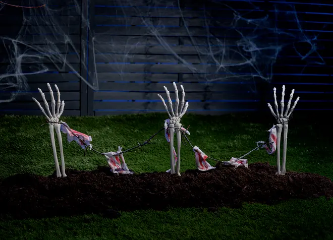 Spöklik Halloween-trädgårdsdekoration med tre skelett-händer på pinnar, sammanlänkade av en trasig girlang, placerad i mörk jord med spindelväv och staket.