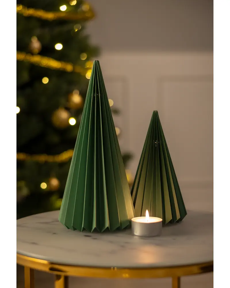 Two green, cone-shaped decorations made from folded paper are displayed on a round marble table. The decorations have a pleated design, giving them a textured, geometric appearance. One is taller and the other is shorter. A small lit tealight candle sits in front of them, adding a warm glow. The background features a blurred Christmas tree with gold ornaments and lights.