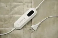 Kontroll till elektrisk filt med ström- och temperaturknappar, digital display och europeisk kontakt, placerad på mjukt, quiltat beige tyg.