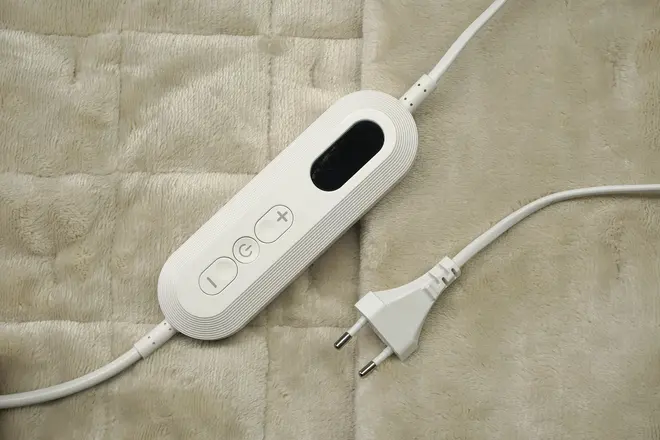 Elektrisk tæppe-controller med tænd/sluk- og temperaturknapper, digitalt display og europæisk stik, placeret på blødt, quiltet beige stof.