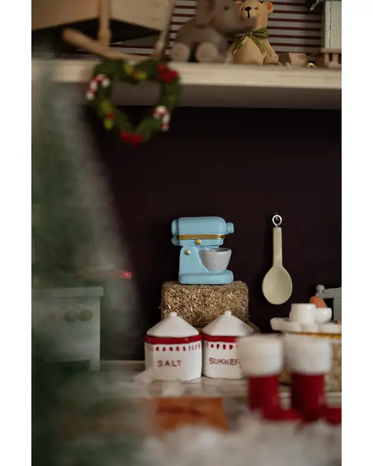 Miniature blå røremaskine med hvid skål, ved siden af keramiske beholdere mærket 'Salt' og 'Sukker', samt en træske. Dekorativt køkkenscene med festlige detaljer.