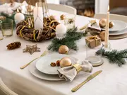 Elegant juleborddækning med guldbestik, hvide tallerkener, linnedserviet med guldring, glas, lys, grangrene, guldkugler og festlig dekoration på hvid dug.