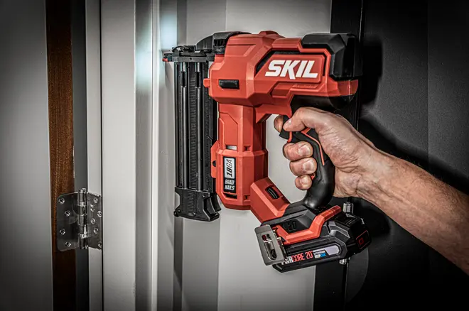 Rød og sort SKIL batteridrevet sømpistol med ergonomisk greb og monteret batteri, brugt til at skyde søm i en ramme.