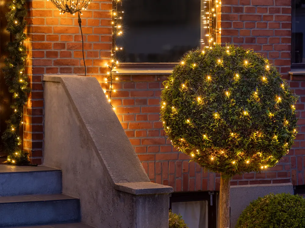 Utomhus ljusslinga med varmvita LED-lampor, lindad runt en klotformad buske och husfasad. Perfekt för festlig eller juldekoration, väderbeständig för trädgård eller uteplats.