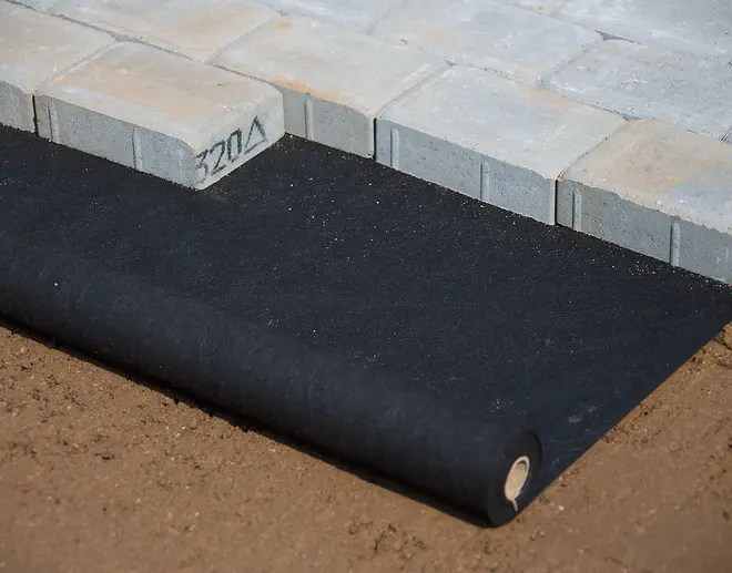 Svart markduk på rulle, delvis utrullad på jord under grå betongplattor, används som ogräsbarriär och markstabilisering vid utomhusläggning.