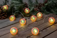 Ljusslinga med runda mosaikglasbollar, flerfärgade plattor och varm gul glöd. Dekorativ, passar för fest eller högtid, på träyta med grankvistar och kottar.