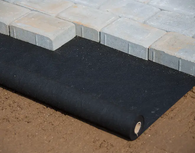 Svart markduk rullas ut under grå marksten på jord, används för ogräskontroll och stabilisering vid utomhusanläggning.