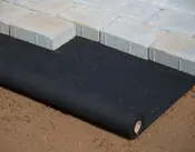 Svart markduk rullas ut under grå marksten på jord, används för ogräskontroll och stabilisering vid utomhusanläggning.