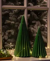 Two decorative cone-shaped objects made from folded green paper, resembling stylized Christmas trees. The paper is pleated vertically, creating a geometric texture. The bases are circular and the tops are pointed. They are placed on a windowsill, surrounded by warm white string lights and festive ornaments, with a snowy tree visible outside the window.