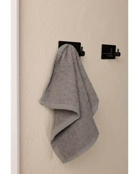 Gråt håndklæde hænger på en moderne sort vægkrog, tekstureret beige væg. Håndklædet ser blødt og absorberende ud, ideelt til badeværelse eller køkken. Minimalistisk design.