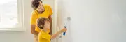 Barn maler en hvid væg med en lille malerrulle, overvåget af en voksen. Begge har gule trøjer på. Rummet er lyst med naturligt lys fra vinduet.