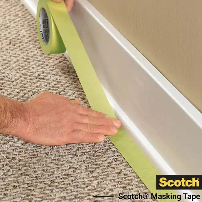 Grøn Scotch afdækningstape påføres langs kanten af et tæppe og en fodliste, ideel til beskyttelse under maling eller renovering. Rullen er nem at håndtere og rive over.