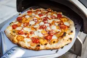 Træfyret pizza med sprød, let brændt skorpe, toppet med smeltet ost, cherrytomater, rødløg og rejer, vist på en metal pizzaspade foran en ovn.