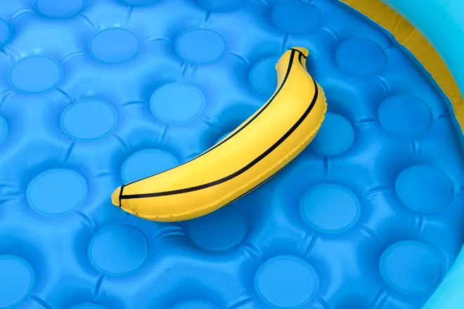 Oppustelig bananformet baderinge, gul med sorte detaljer, til brug i pool. Ligger på blå oppustelig pools overflade. Sjovt og unikt pooltilbehør til børn eller voksne.