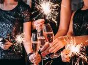 Tre kvinnor i svarta paljettklänningar håller tomtebloss och skålar med champagneglas, vilket skapar en festlig stämning för fester eller speciella tillfällen.