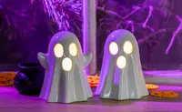 Two glossy white ceramic ghost-shaped decorations with illuminated eyes and mouth openings, creating a glowing effect. Each ghost has a smooth, rounded surface and slightly raised arms, giving a playful and friendly appearance. The lights inside the ghosts emit a warm yellow glow. The decorations are placed on a wooden surface with Halloween-themed accents in the background.