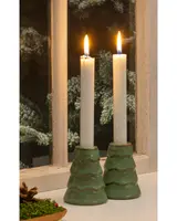 Two tall white candles are placed in ceramic holders shaped like stylized green Christmas trees with a glossy finish. The holders have a tiered design with wavy ridges, and the candles are lit, casting a warm glow. The scene is set on a windowsill with a snowy pine tree visible outside, creating a cozy, festive atmosphere.