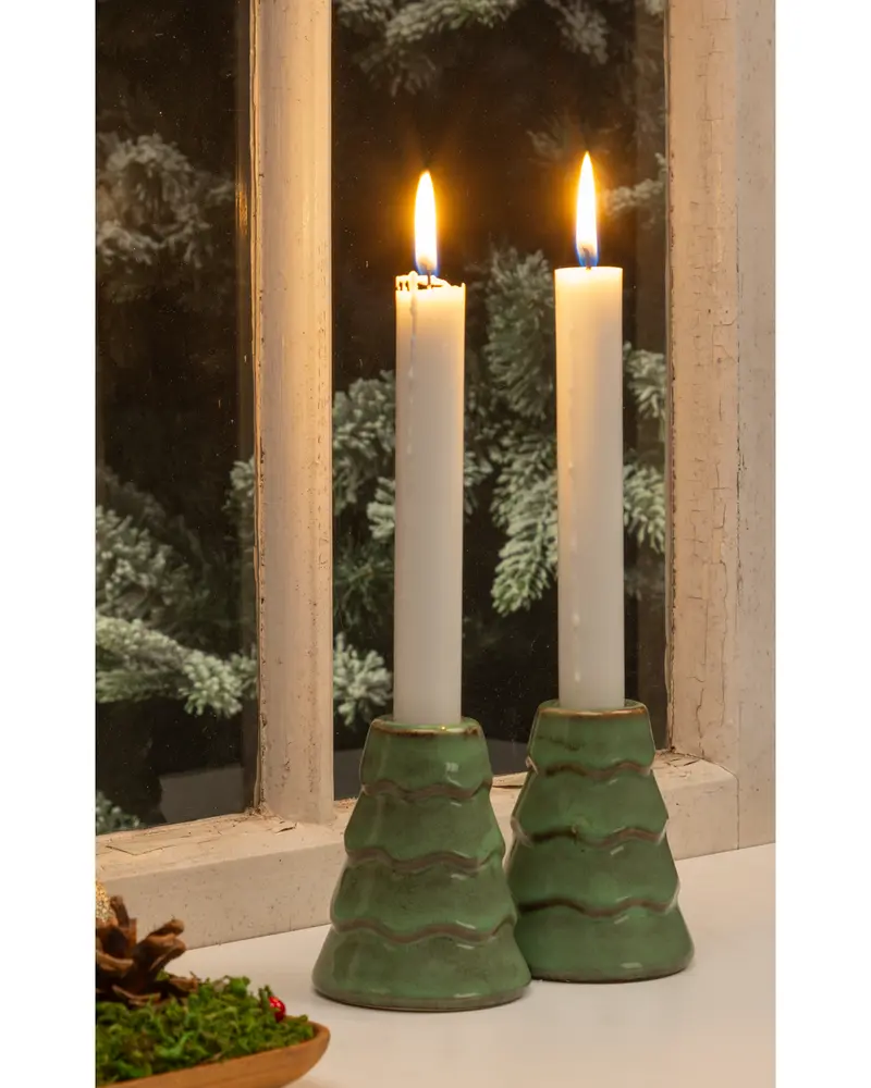Two tall white candles are placed in ceramic holders shaped like stylized green Christmas trees with a glossy finish. The holders have a tiered design with wavy ridges, and the candles are lit, casting a warm glow. The scene is set on a windowsill with a snowy pine tree visible outside, creating a cozy, festive atmosphere.