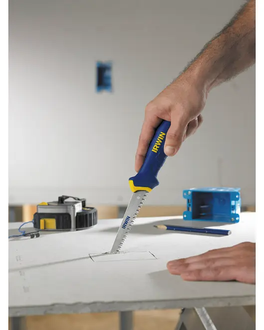 Handsåg med blått och gult handtag, märkt IRWIN, sågar ett rektangulärt hål i gipsskiva. Vass, spetsig klinga för exakta snitt. Måttband och penna intill.