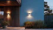 Moderne udendørs væglampe med cylindrisk sort og hvidt design, monteret på husets facade, udsender varmt lys opad og nedad, ideel til moderne indgange eller terrasser.