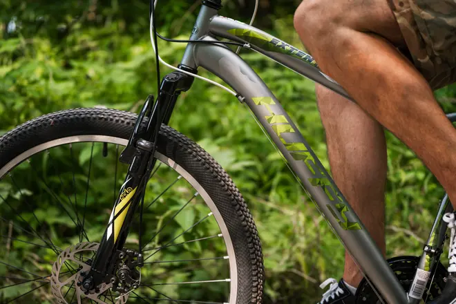 Närbild på silverfärgad mountainbike med framdämpare, grova däck, skivbromsar och grönt Hurley-märke. Cyklist i shorts delvis synlig, utomhus på stig.